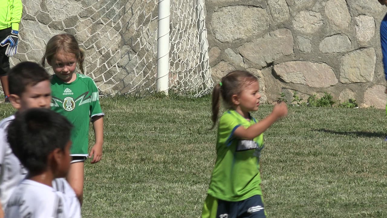 A girl in a green soccer jersey stands by the goal net while other children pass through the foreground on the San Jose Del Cabo grass field.