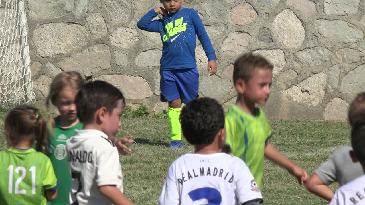 Children in green, white, and blue soccer uniforms gather on a grass field in San Jose Del Cabo, Mexico, with a goal net and stone wall behind them.
