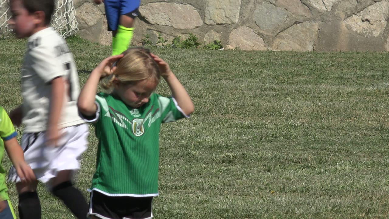 A blonde girl in a green soccer jersey stands near the center of a grass field in San Jose del Cabo, Mexico, while children in white uniforms blur past her.