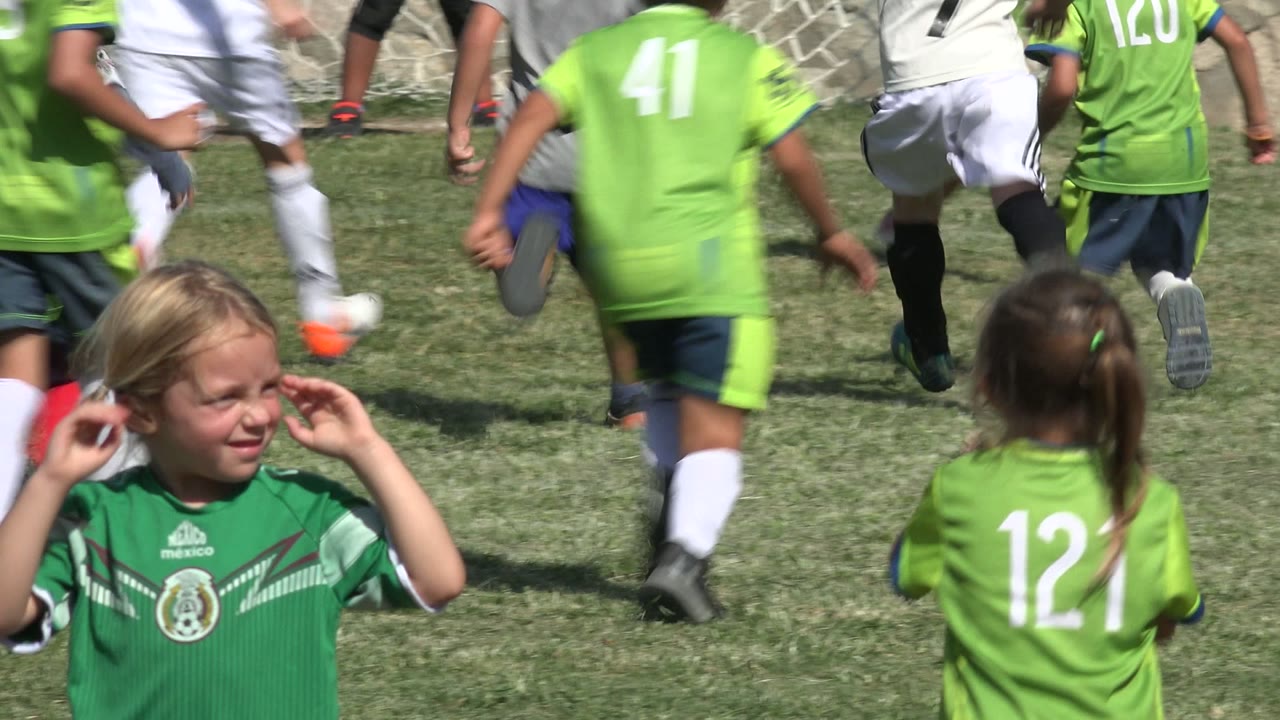 Young girl in green soccer jersey with children running behind her on grass.