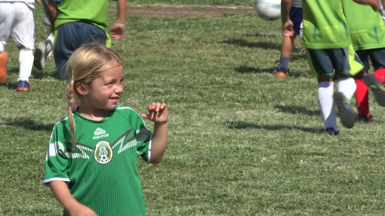 At 0:01 in San Jose del Cabo, Mexico, the girl is centered in the foreground wearing a green jersey while a group of children runs upfield in the background.