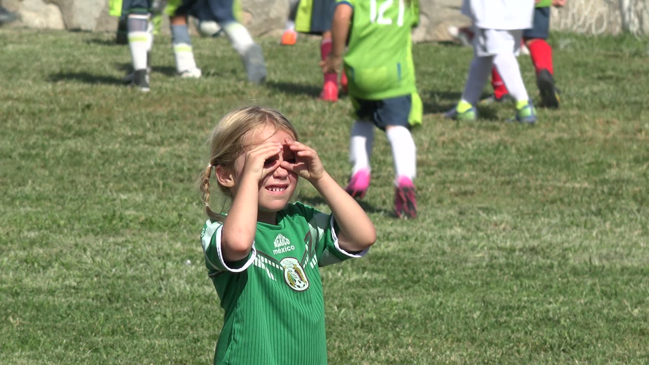 Young girl in a green soccer jersey shading her eyes on a grass field.