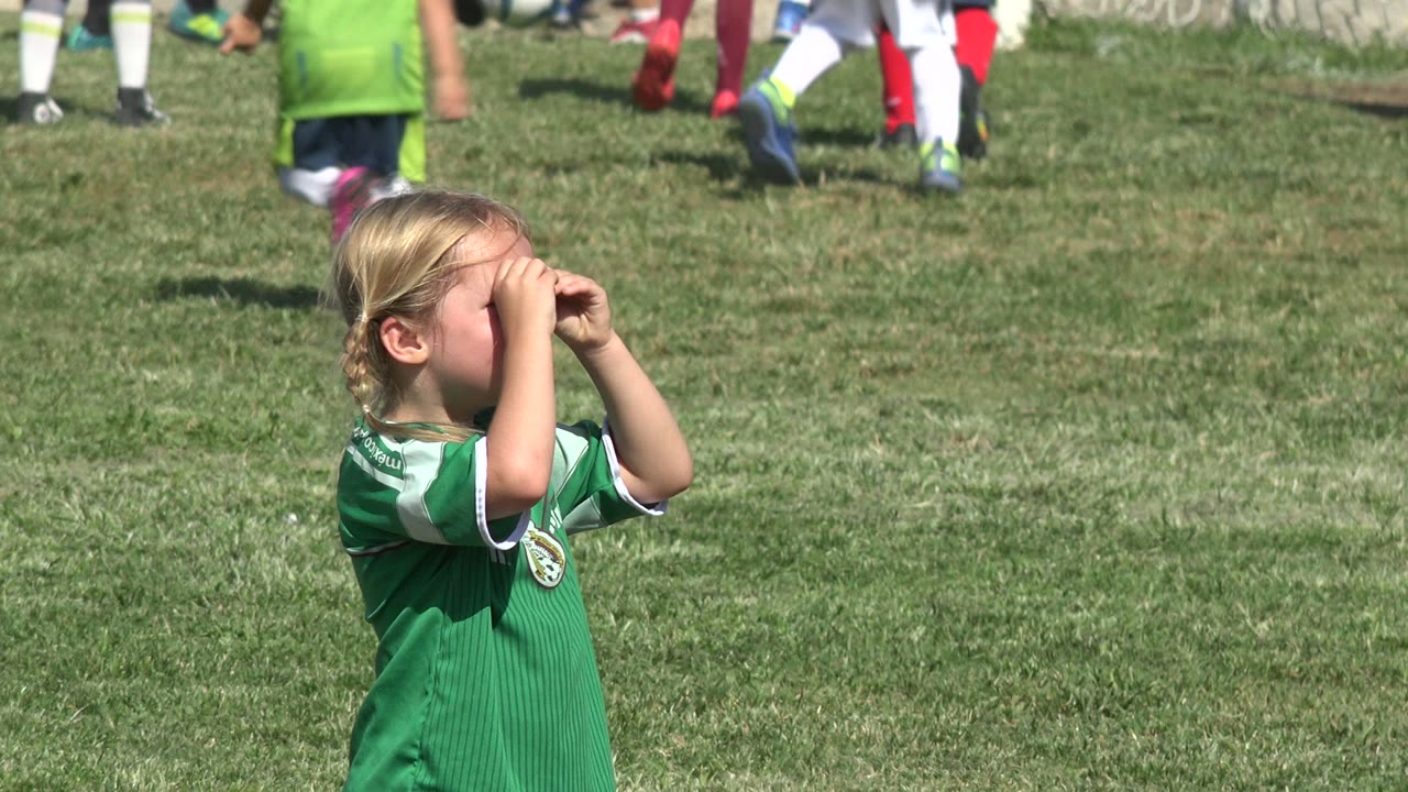 The girl turns slightly in profile with her hands at her brow, looking across the grass as children in green and white uniforms pass through the background in San Jose del Cabo, Mexico.