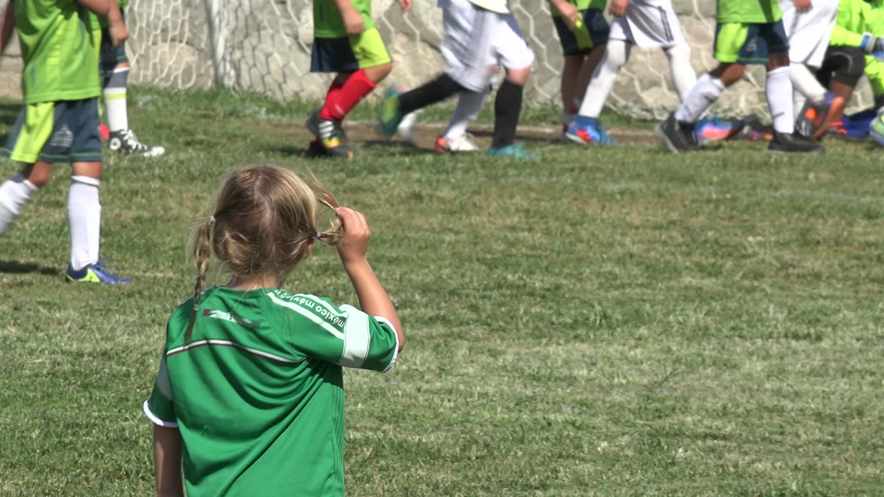 Young girl in a green jersey watching children on a soccer field.