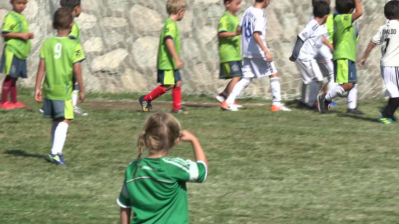 Children in green and white soccer kits walk along the grass near netting and a stone wall, with the watching girl still visible low in the foreground in San Jose Del Cabo, Mexico.