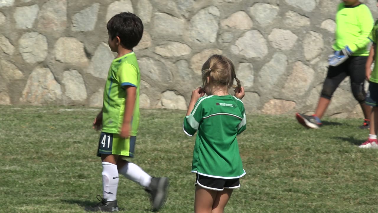 A blonde girl in a green jersey walks away from the camera on the grass in San Jose Del Cabo, Mexico, while a boy in a green soccer uniform stands near the stone wall.