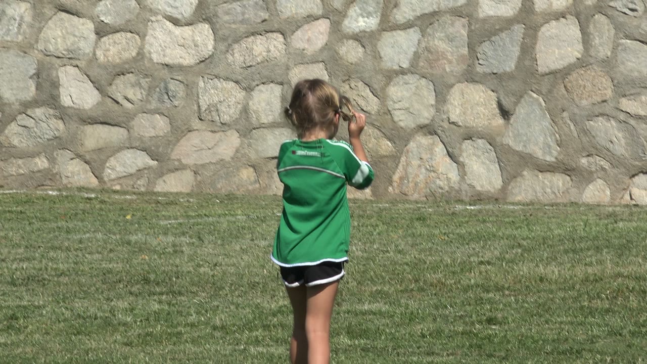 Girl in a green soccer jersey on a grass field near a stone wall.
