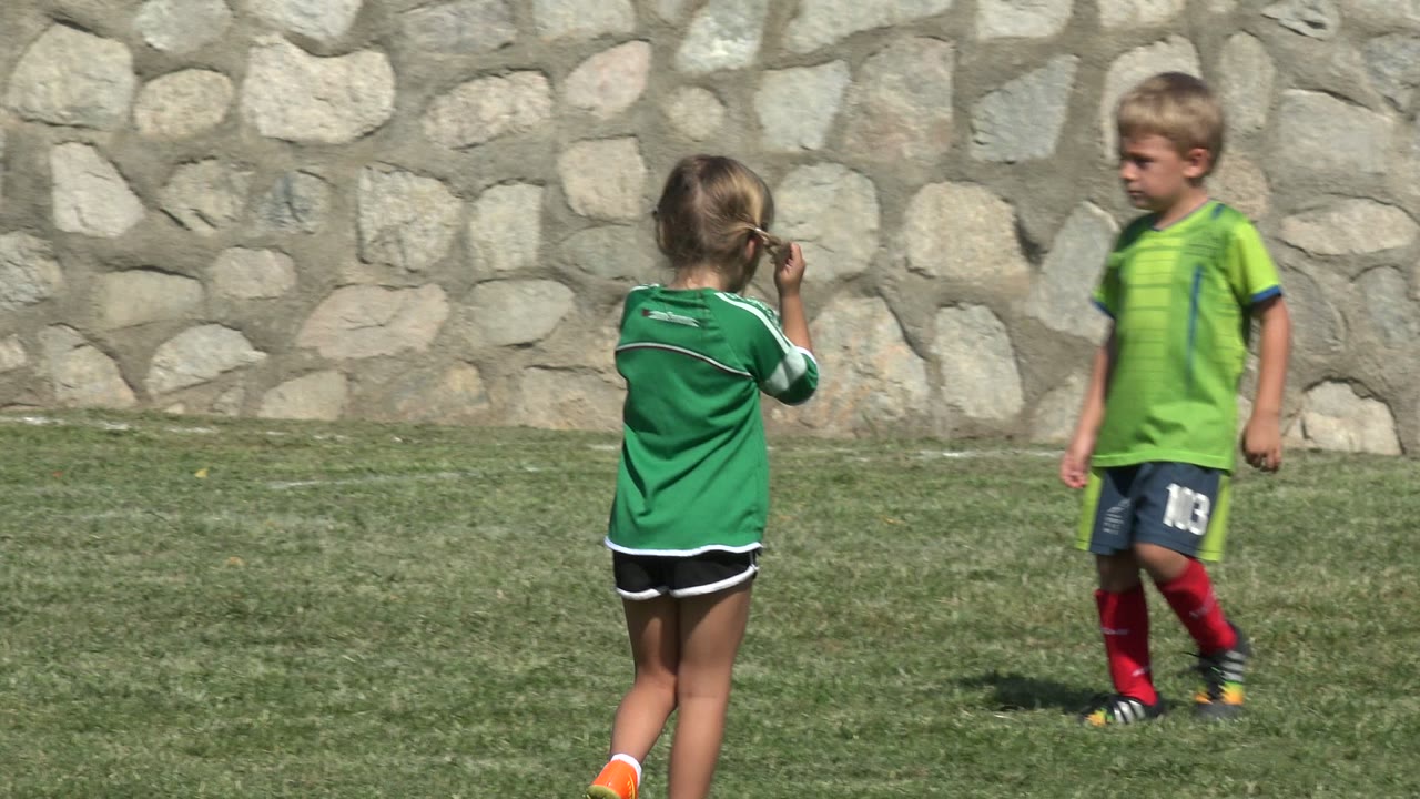 A girl in a green jersey stands near the left side of the frame while a boy in a bright green soccer shirt walks across the grass in San Jose Del Cabo, Mexico.