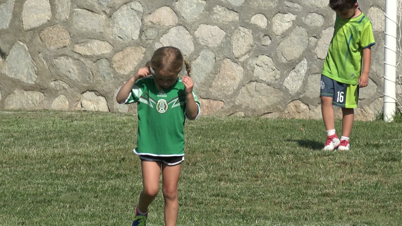 The girl holds the collar of her green soccer jersey while standing on the field in San Jose del Cabo, Mexico, with a white goal post and another child in the background.
