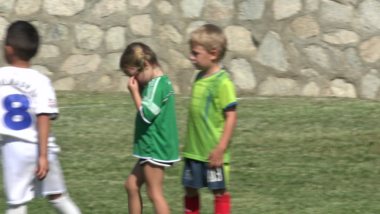 Children in soccer uniforms standing on grass near a stone wall.