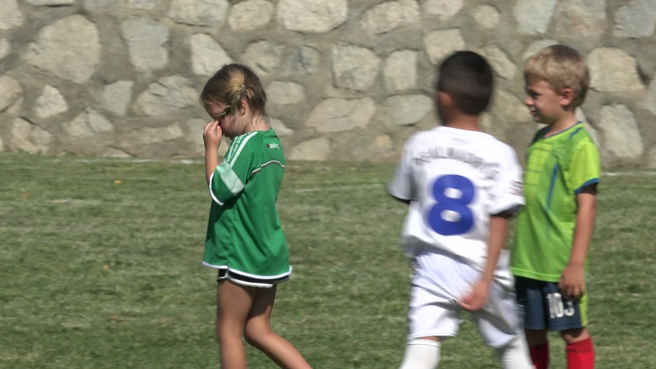 A girl in a green soccer jersey walks across the grass in San Jose Del Cabo, Mexico, while boys in green and white uniforms move nearby against a stone wall.