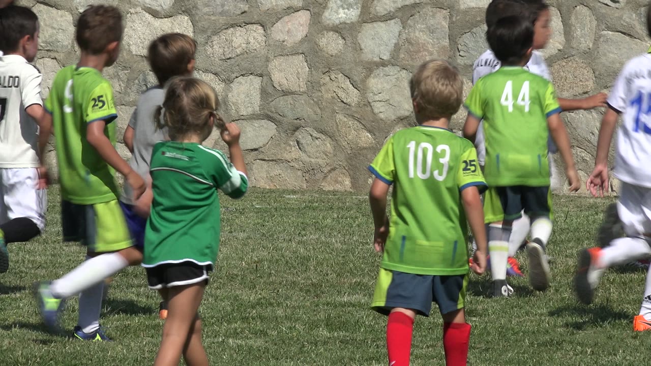 Children in green and white soccer uniforms running on grass near a stone wall.