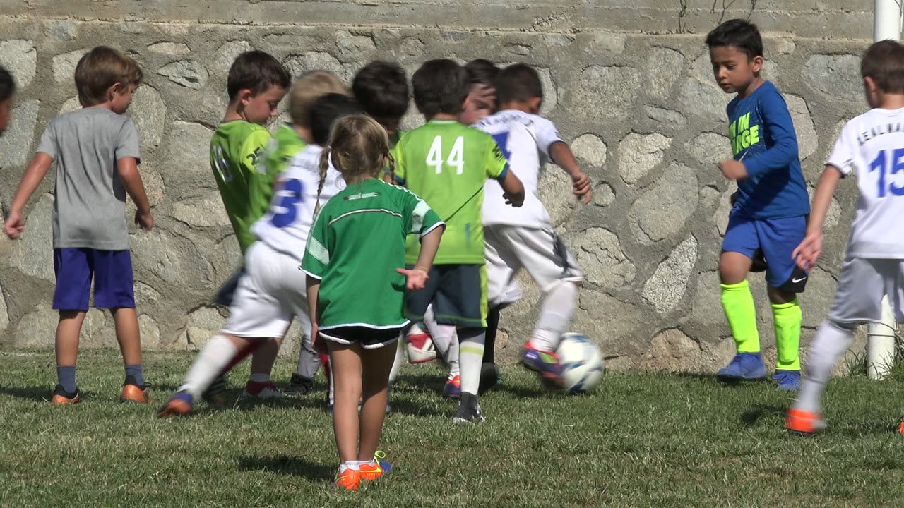 A cluster of children in green and white uniforms move around the ball beside the goal net in San Jose del Cabo, Mexico, with a child in a gray shirt standing nearby.