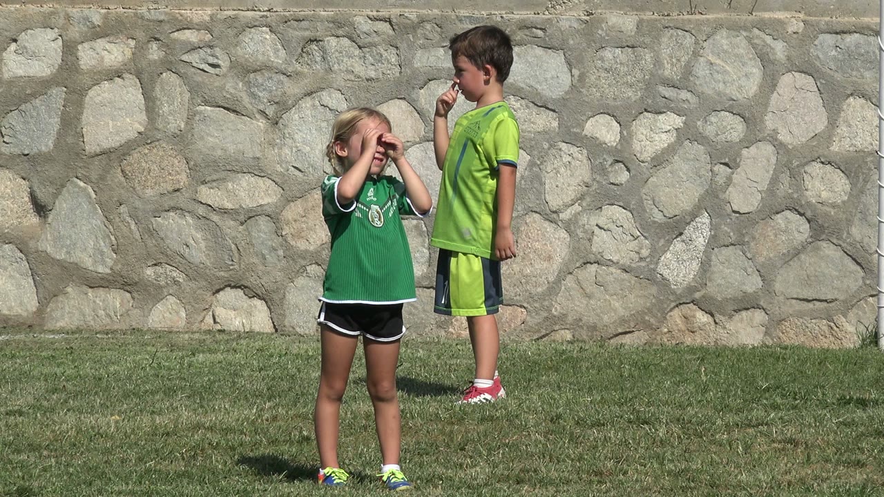 A young blonde girl covers her face in the foreground while a boy in a green shirt stands farther back on the soccer field in San Jose del Cabo, Mexico.