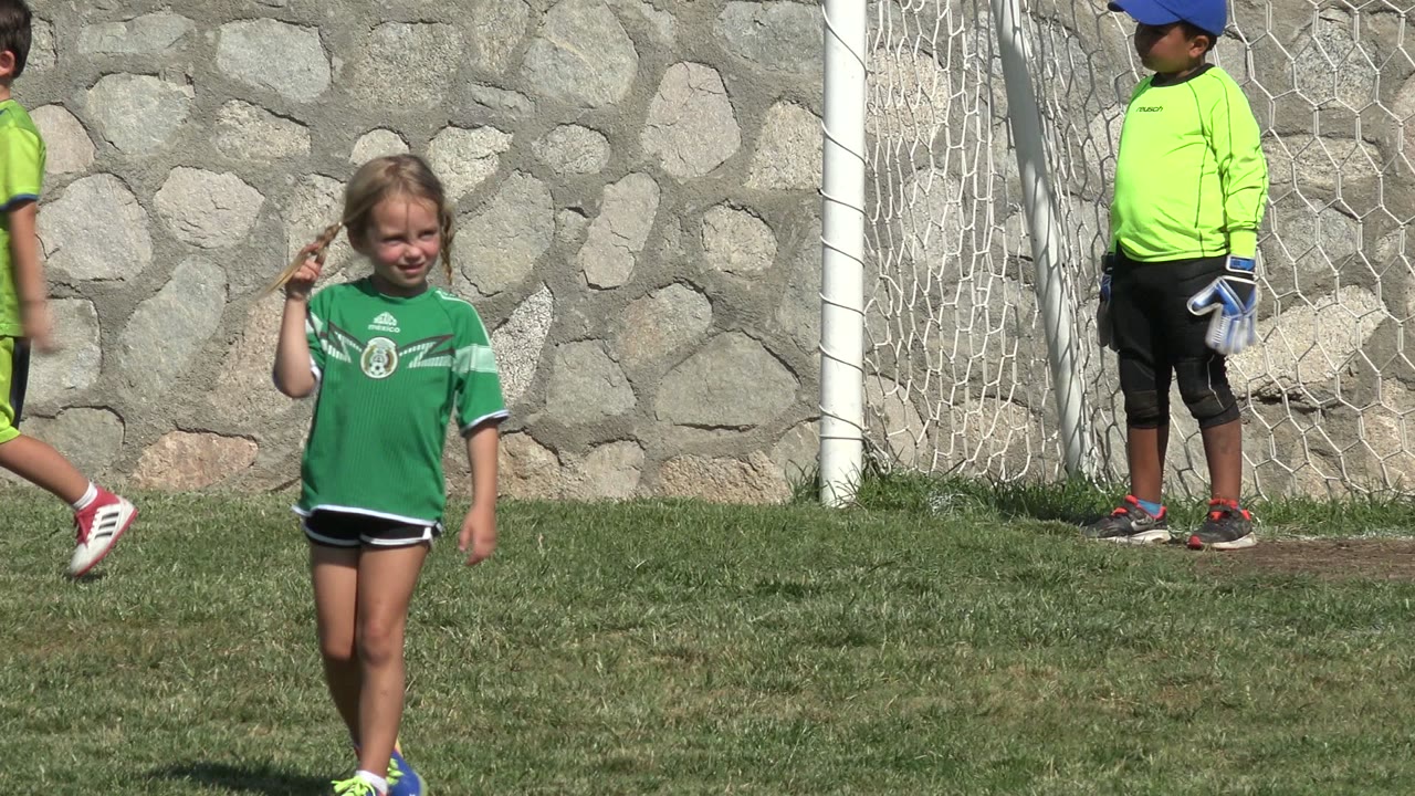 The girl in a green soccer shirt faces the camera on the San Jose del Cabo field, with a white goal net at the side and a child goalkeeper behind her.