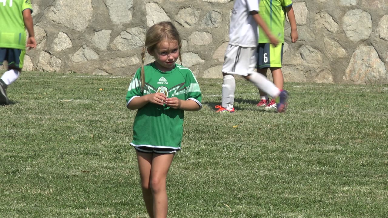 Young girl in a green soccer jersey stands on a grass field in San Jose del Cabo, Mexico, with other children in soccer uniforms crossing behind her near a stone wall.