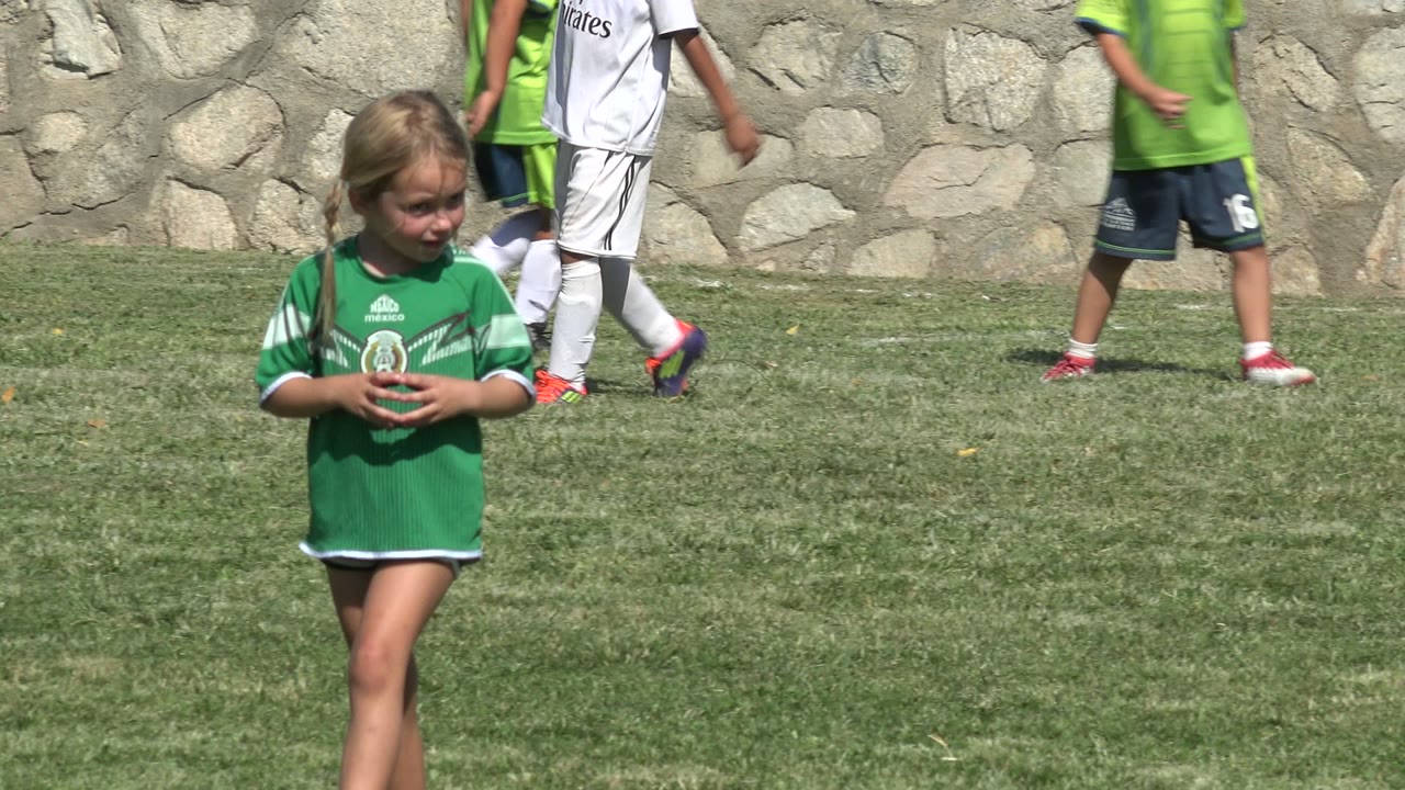 Girl in a green jersey stands near the center of the frame in San Jose del Cabo, Mexico, holding her hands while children in green and white soccer kits move across the background.