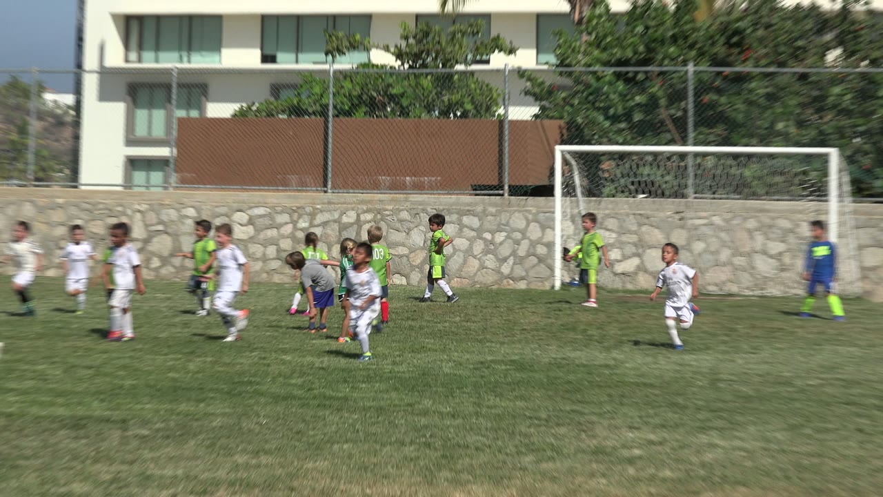 Children in green, white, and gray soccer clothing cluster near the goal area in San Jose del Cabo, Mexico, with the stone wall and modern buildings in the background.