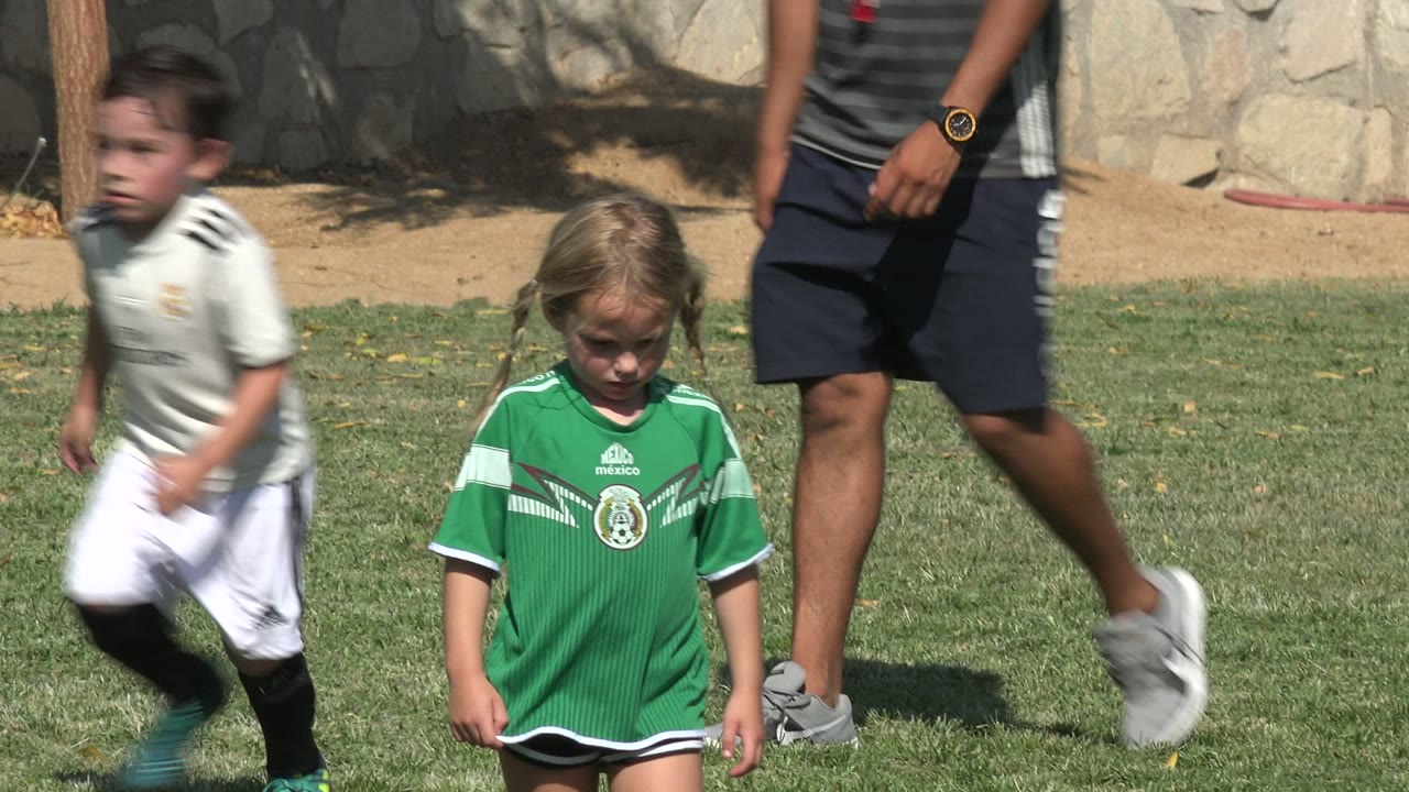 Young children on a grass soccer field with adult legs in the background.