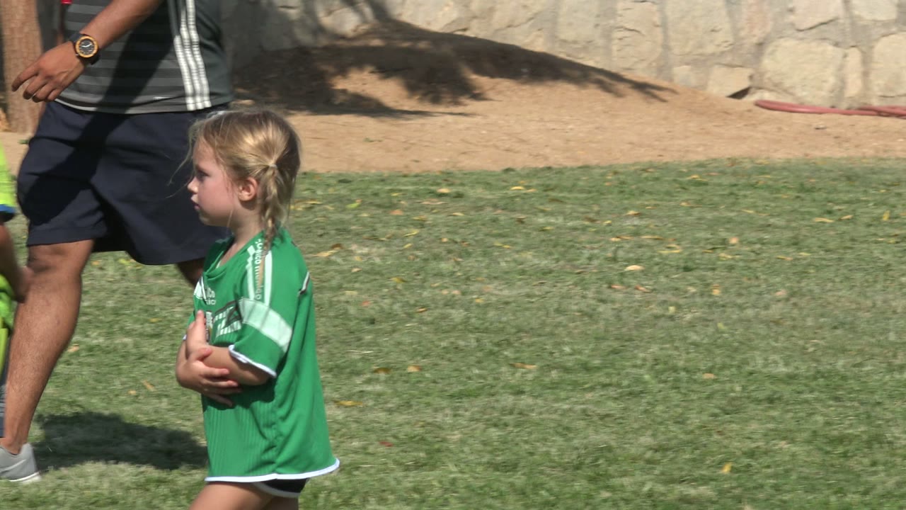 A young girl in a green jersey walks in profile across the San Jose del Cabo grass field, with adult coach legs and the stone wall visible behind her.