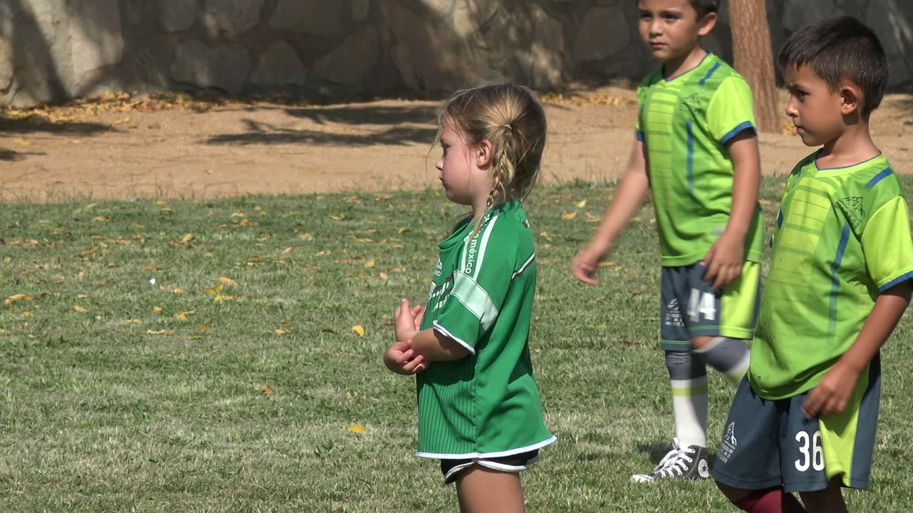 In San Jose del Cabo, Mexico, a girl and a boy in green soccer uniforms stand in profile on a sunny grass field with a stone wall behind them.