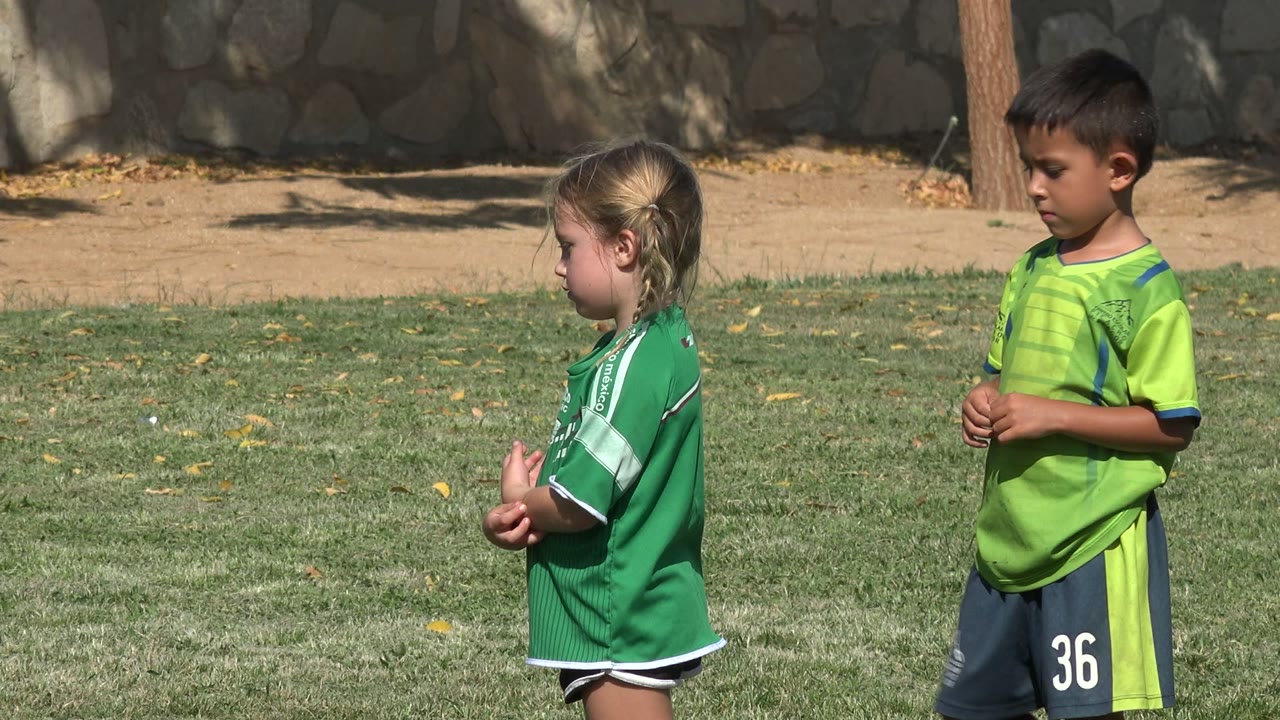 Blonde girl in a green soccer jersey standing on grass near a boy in lime green uniform.