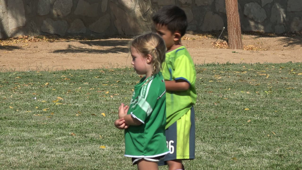 In San Jose del Cabo, Mexico, the girl remains in profile in her green jersey while the boy in lime green shifts behind her against the same grass and stone wall background.