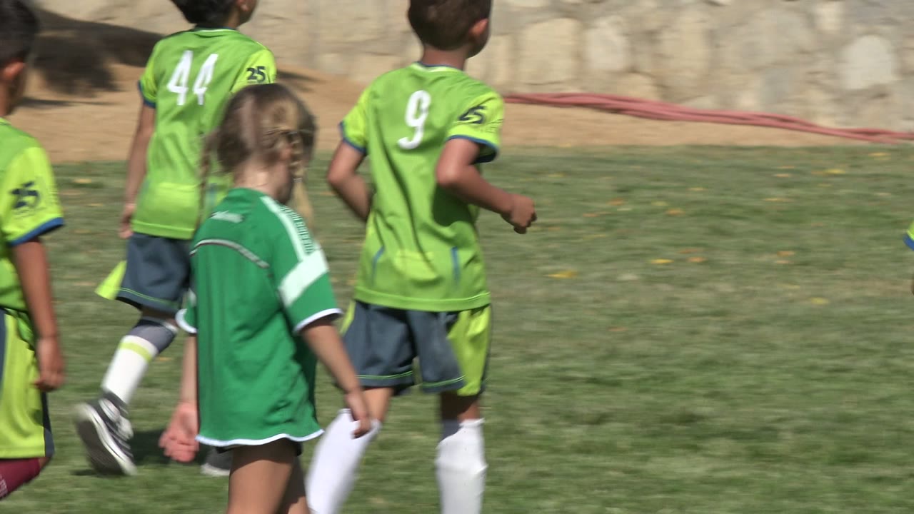 Children in green soccer uniforms run on a grass field as a young girl stands nearby.