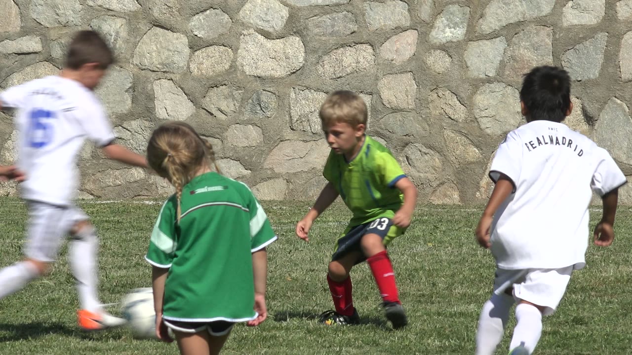 Children in soccer uniforms play on a grass field beside a stone wall.