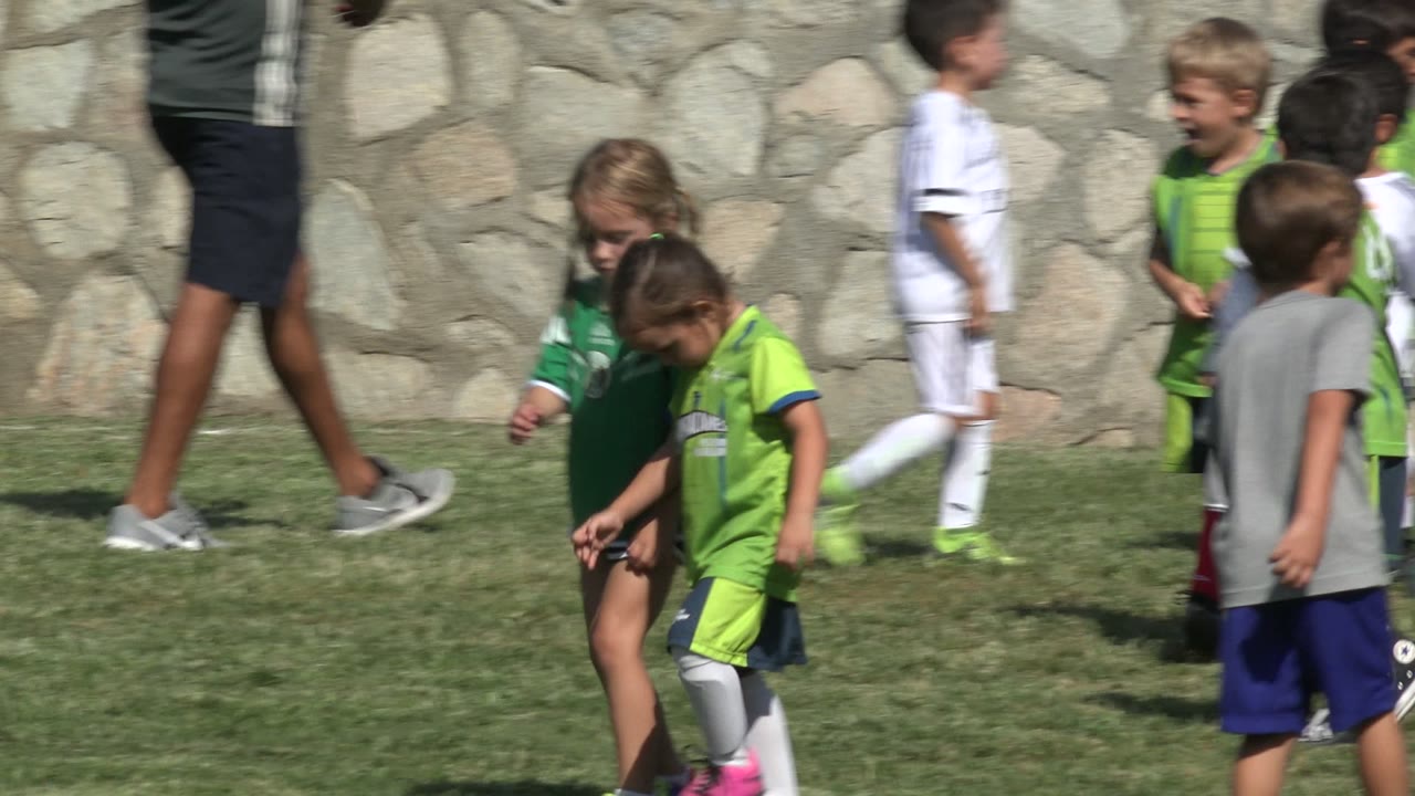 Children in green soccer uniforms walking on grass near a stone wall.