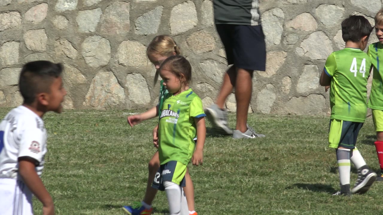 Young players in green jerseys gather and walk on the grass in San Jose del Cabo, Mexico, while a child in white passes through the foreground.