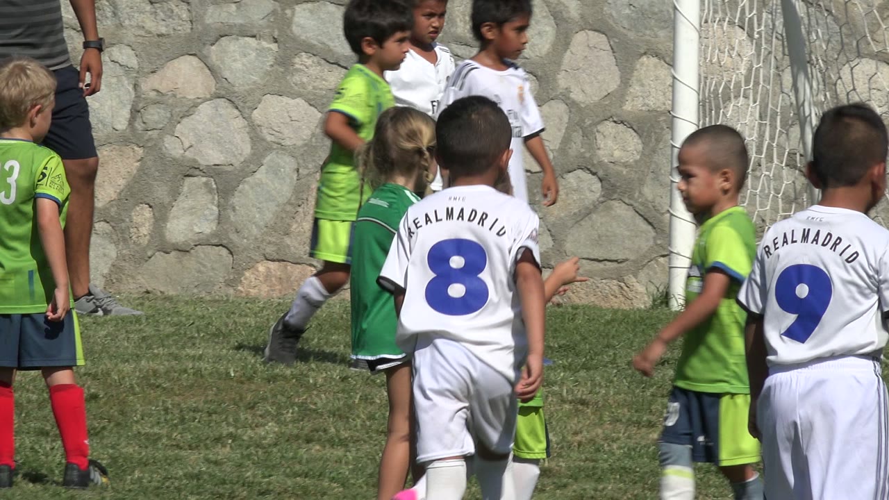 Several children in white and green soccer uniforms stand and move in front of the goal net in San Jose del Cabo, with jersey numbers visible from behind.