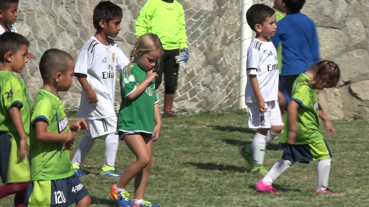 A cluster of young soccer players in green, white, and bright goalkeeper shirts stands and walks near the goal net on a sunlit field in San Jose del Cabo, Mexico.