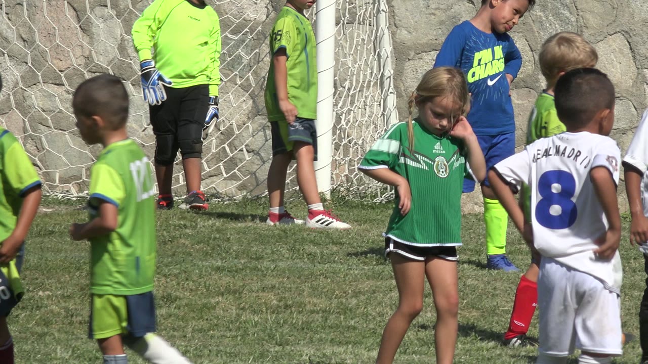 Kids in white, green, and blue soccer kits stand near the goal area on sunlit grass in San Jose del Cabo, Mexico.