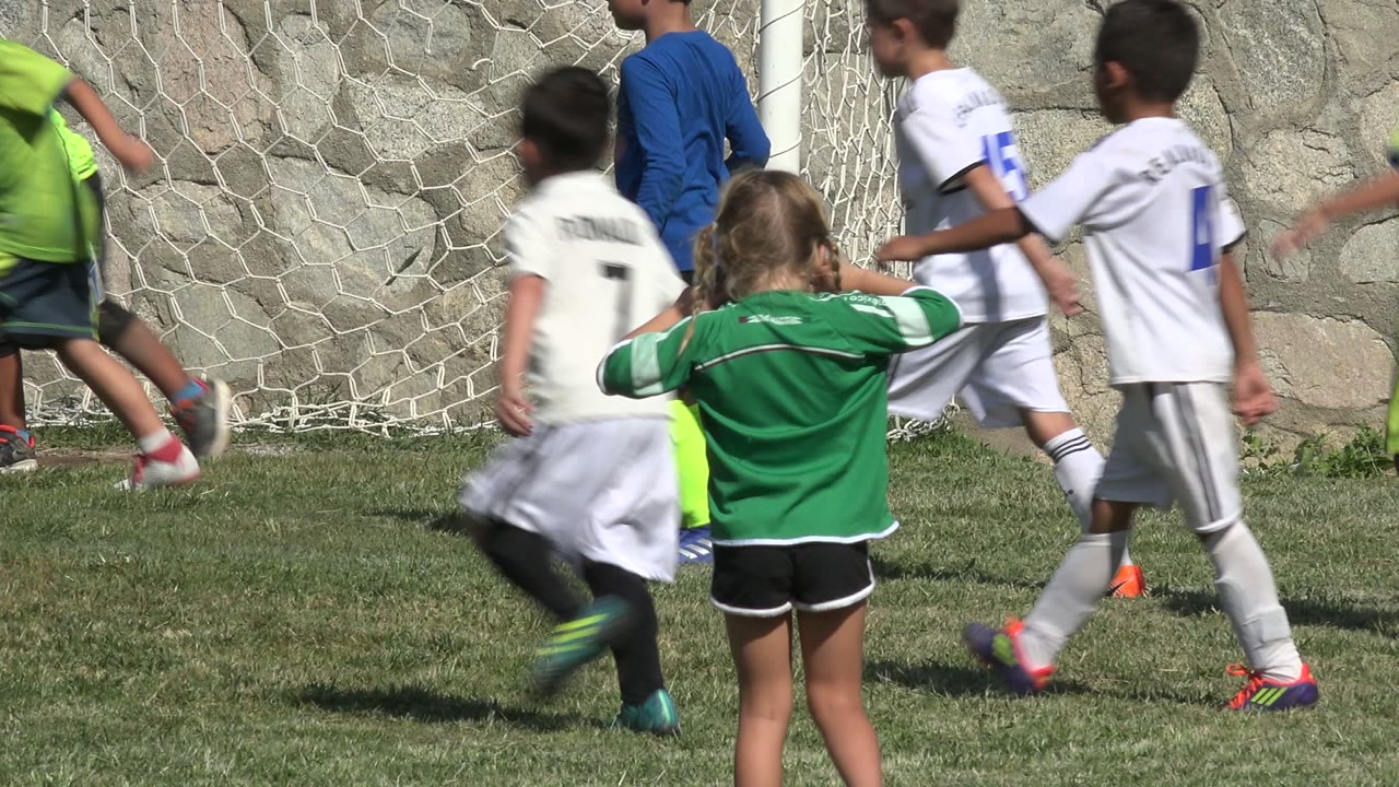 A blonde girl in a green soccer jersey stands near the foreground while children in white uniforms pass the goal net in San Jose del Cabo, Mexico.
