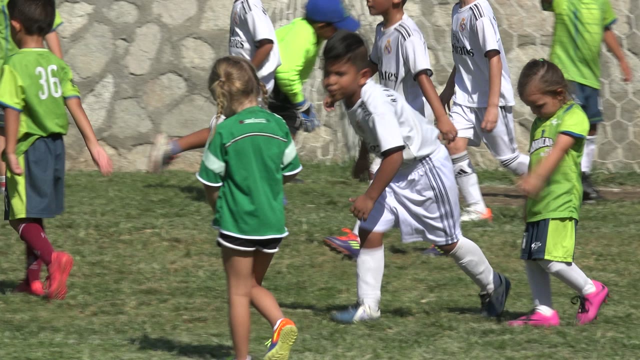 Children in green and white soccer uniforms play on a grass field in San Jose del Cabo, Mexico, with a girl in green seen from behind in the foreground and a goal net at the right.
