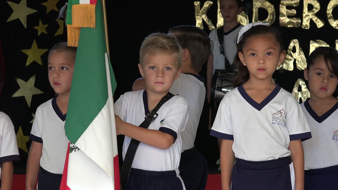 Children in white shirts stand beside a green, white, and red flag indoors.