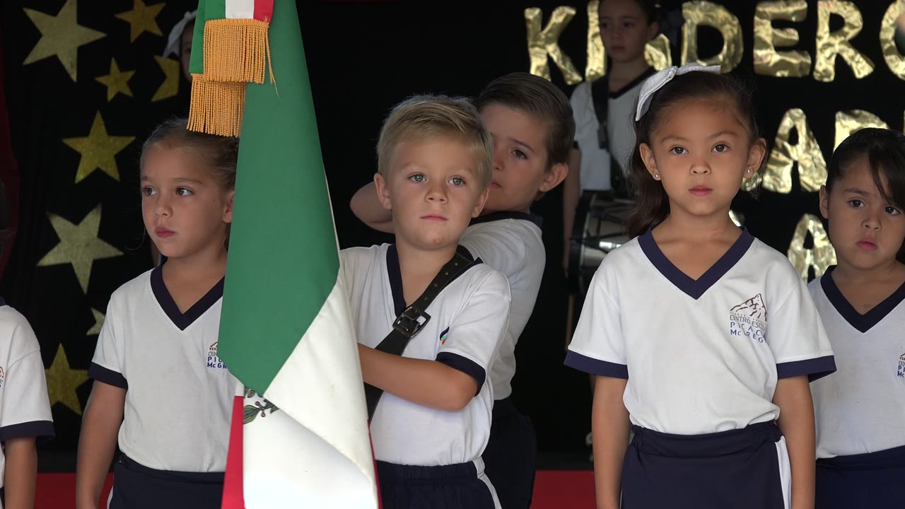 At 0:01 in San Jose del Cabo, Mexico, the boy holding the flag faces forward as other children in matching white shirts stand closely behind him.