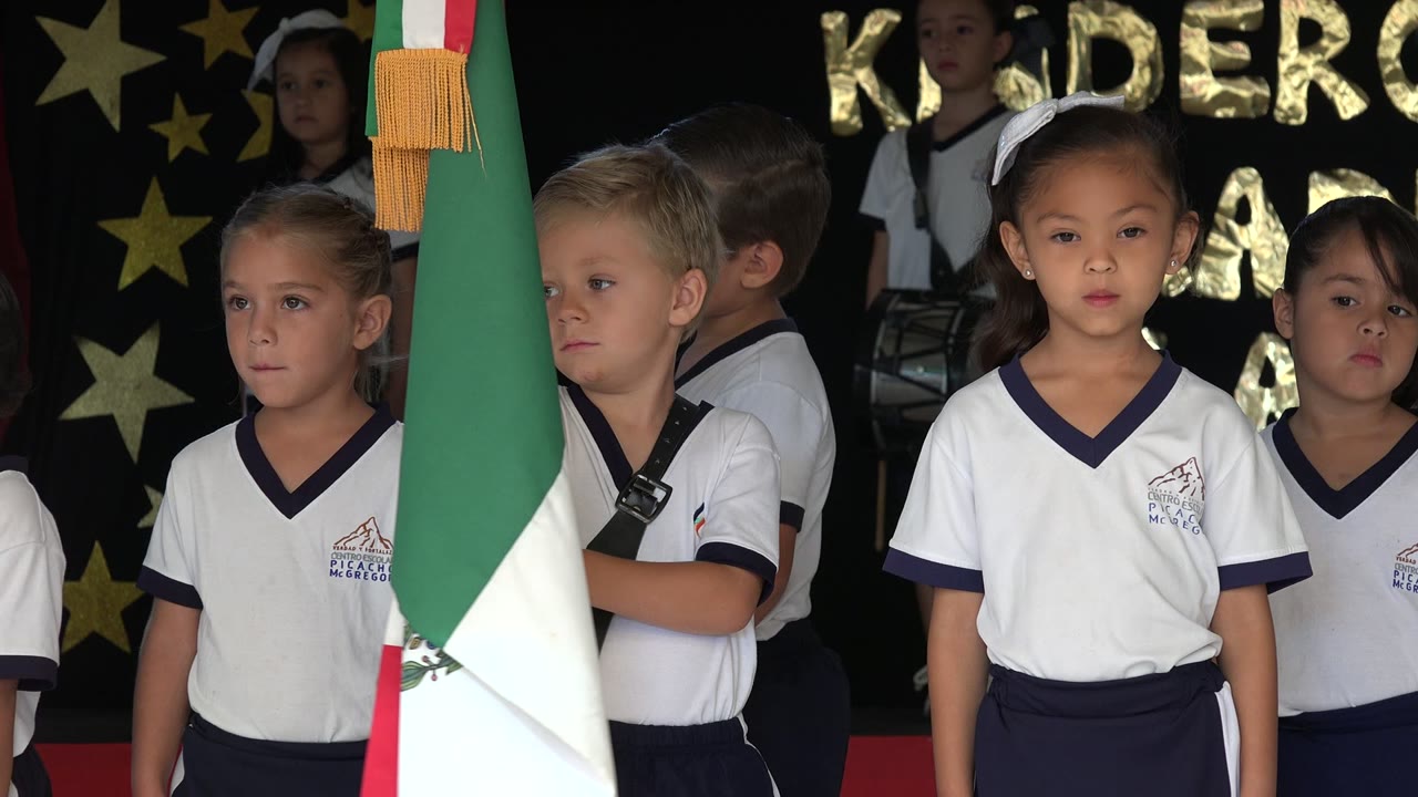 Children in school uniforms stand on a decorated stage beside a Mexican flag.