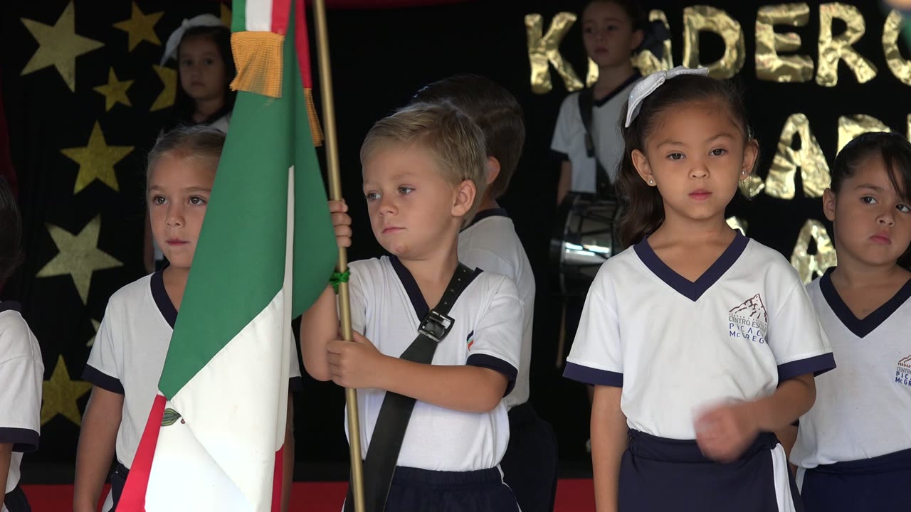 At 0:01, a girl in a white uniform faces the camera while other children stand nearby under gold star decorations and partial graduation lettering.