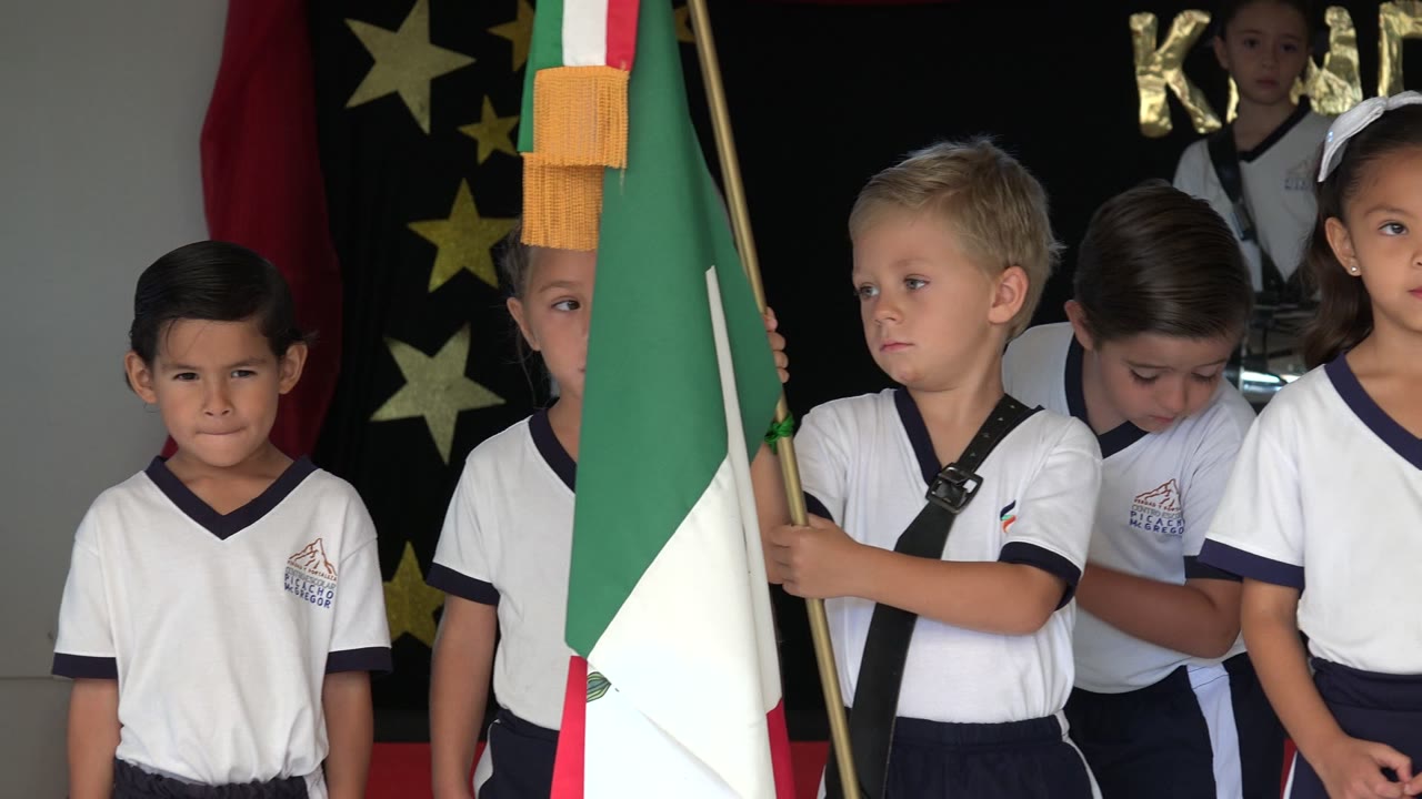 Young schoolchildren in white uniforms stand in San Jose Del Cabo, Mexico beside a large green, white, and red flag, with a dark stage backdrop and gold stars behind them.