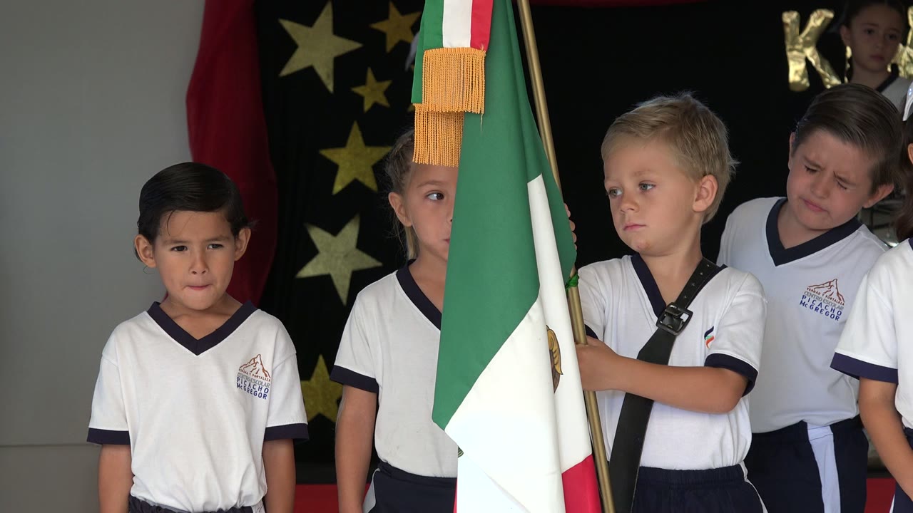 Children in white uniforms wait in San Jose Del Cabo, Mexico while a foreground flag partially blocks one child and other students gather behind the front row.