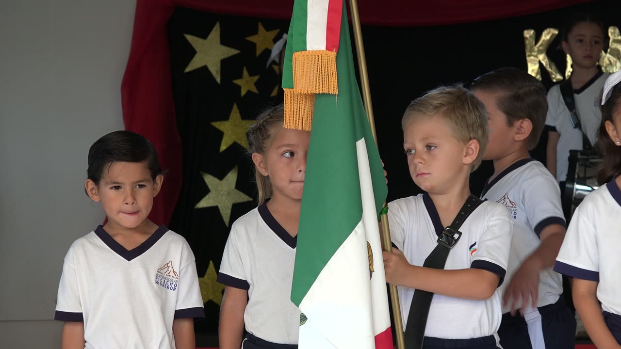 A line of children in white uniforms stands beside a flag and stage decorations.