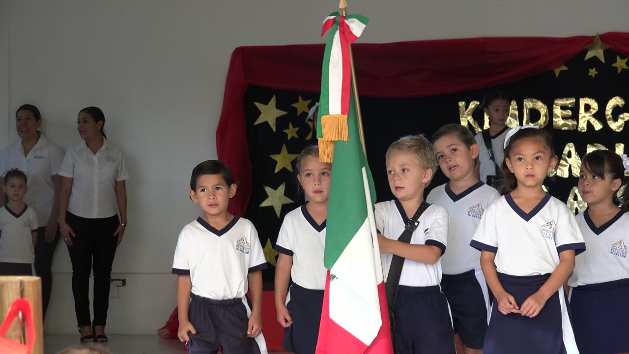 Children in white uniforms stand on an indoor stage in San Jose del Cabo, Mexico, beside a Mexican flag while adults stand behind them near a decorated backdrop.