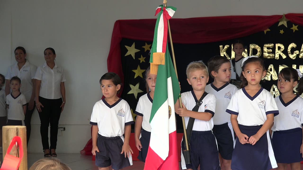 The children remain gathered around the Mexican flag on the San Jose del Cabo stage, with several faces turned toward the audience and teachers visible in the background.