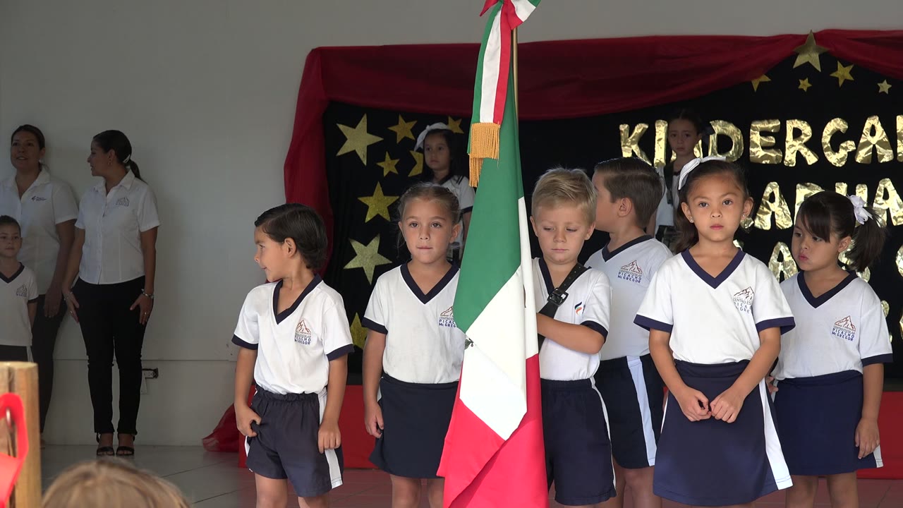 Children in uniforms stand near a Mexican flag on an indoor stage.