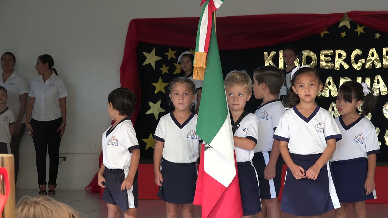 A group of children in uniforms waits in San Jose del Cabo, Mexico, while one child looks toward the side and the Mexican flag hangs between the camera and the group.