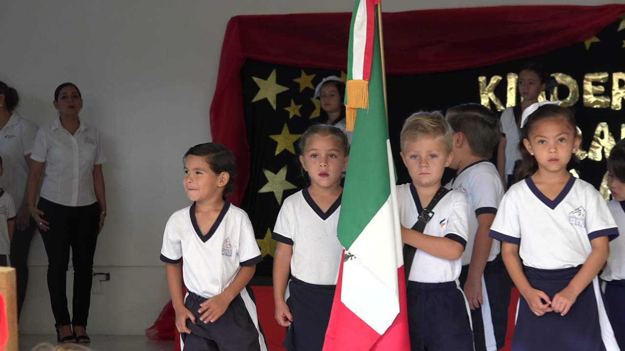 Children in white uniforms stand on a small stage near a Mexican flag.