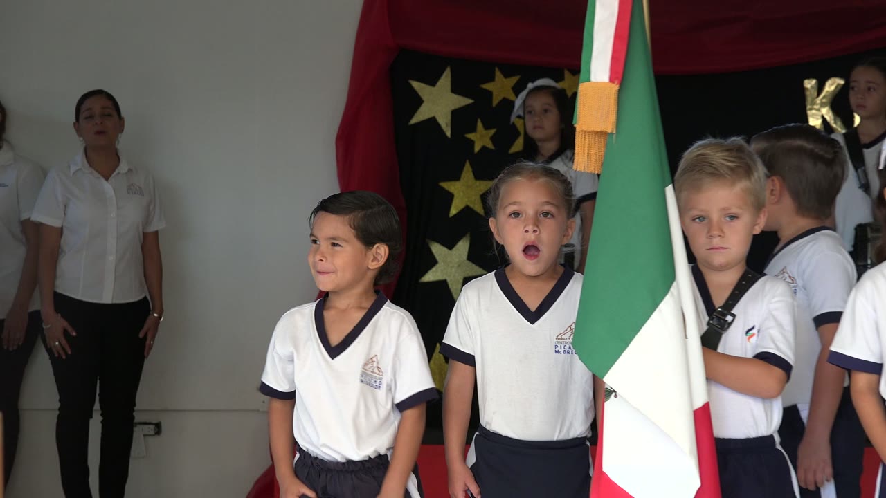 Mid-wide view of children on the stage in San Jose del Cabo, Mexico, with the Mexican flag partially covering the center-right foreground and a dark star backdrop behind them.