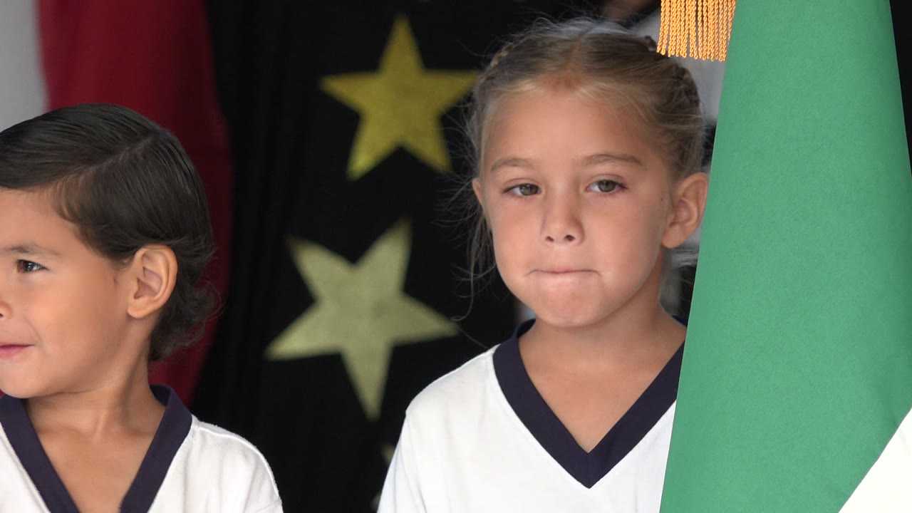 Two children in white uniforms stand close to a green and white flag during a ceremony preparation moment in San Jose del Cabo, Mexico.